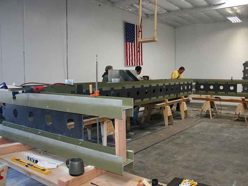 Rock West's low dielectric large antenna test fixture is show under construction in a manufacturing bay with an American flag on the wall. Three technicians are working along the length of the fixture, which is on its side and elevated on wood sawhorses.